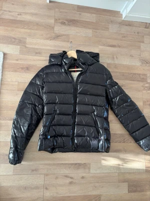 Svart dunjacka från Moncler - Snygg svart dunjacka från Moncler med glansig finish och klassisk quiltad design. Jackan har huva, två dragkedjeförsedda fickor och Moncler-logga på ärmen. Perfekt för kalla vinterdagar och ger en stilren look.