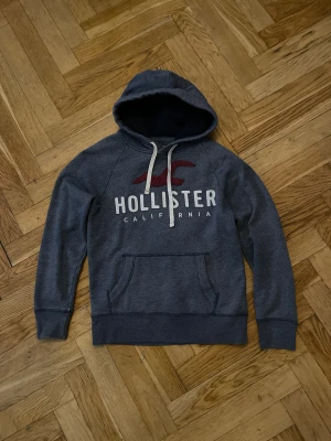 Mörkgrå Hollister hoodie med tryck - Mörkgrå hoodie från Hollister med stor vit logga och röd fågel på bröstet. Klassisk känguruficka framtill och vita dragsnören i huvan. Mjuk bomullsblandning som känns skön mot huden. Perfekt för chill dagar eller när du vill ha en avslappnad look.