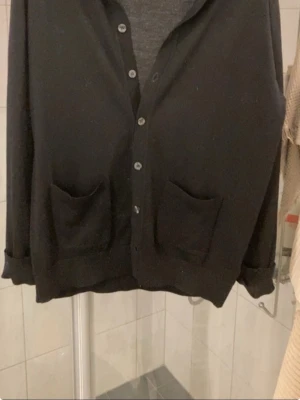 Svart basic kofta med fickor - En enkel svart kofta från Oscar Jacobson med knappar framtill och två rymliga fickor. Koftan har långa ärmar och en avslappnad passform, perfekt att slänga över en t-shirt. Materialetär mjukt och bekvämt, och modellen är klassisk utan synligt märke. Perfekt för en daglig outfit. Har ett litet hål baktill som syns på bilderna. Annars är den i topp skick. Priset gör att diskutera. Passar runt 185-190cm