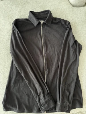 Svart overshirt från Ciszere - Stilren svart overshirt från Ciszere med rak passform och dragkedja framtill. Skjortkrage och långa ärmar med knapp vid manschetten. Tillverkad i ett mjukt bomullstyg som känns bekvämt och passar perfekt till lager-på-lager-stil.