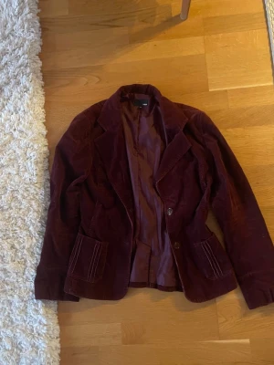 Vinröd manchesterkavaj från H&M - Säljer en vinröd kavaj i manchester från H&M. Jackan har klassisk krage, två stora fickor framtill och stängs med två knappar. Perfekt för dig som vill ha en retro och stilren look.