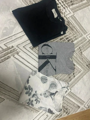 3-pack t-shirts Calvin Klein & mönstrad - Säljer ett 3-pack t-shirts: en svart basic, en grå Calvin Klein med stor logga framtill och en vit t-shirt med blommigt mönster. Alla har rund hals och korta ärmar. Perfekta för en clean och avslappnad stil.
