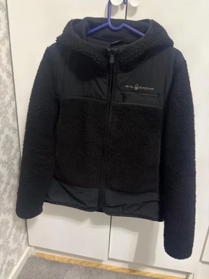 Svart teddyjacka från Sail Racing - Mysig svart teddyjacka från Sail Racing med huva och dragkedja framtill. Jackan har en bröstficka med dragkedja och logga på vänster sida. Materialet är fluffigt teddytyg kombinerat med släta partier för en sportig look. Perfekt för kalla dagar.