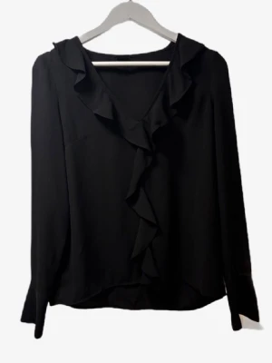 Svart blus med volangdetalj - Stilren svart blus med v-ringning och volangdetaljer längs halsringningen och framtill. Blusen har långa ärmar och är tillverkad i ett lätt, mjukt tyg som ger ett snyggt fall. Perfekt för dig som gillar en chic och enkel look.