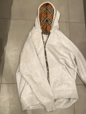 Grå hoodie med Burberry-mönstrad huva - Säljer en grå zip-hoodie från Burberry med klassiskt rutigt foder i huvan. Hoodien har dragkedja framtill och en avslappnad passform. Perfekt för dig som vill ha en stilren men ändå unik look. Materialet känns mjukt och bekvämt. priset går att diskutera. Passar båda m och L
