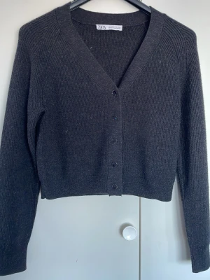 Mörkgråa ribbad kofta från Zara - Stilren mörkgrå ribbad kofta från Zara med v-ringning och knäppning framtill. Koftan har långa ärmar och en croppad passform som ger en modern look. Perfekt att matcha med jeans eller kjol för en enkel men snygg outfit.