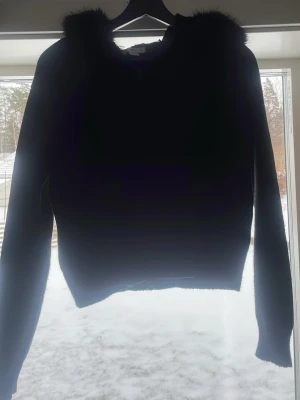 Svart hoodie med fuskpäls - Säljer en svart hoodie med huva som har fuskpäls runt kanten. Hoodien har långa ärmar och en klassisk, enkel design. Perfekt för dig som vill ha en mysig och stilren look. Materialet ser ut att vara mjukt och bekvämt.