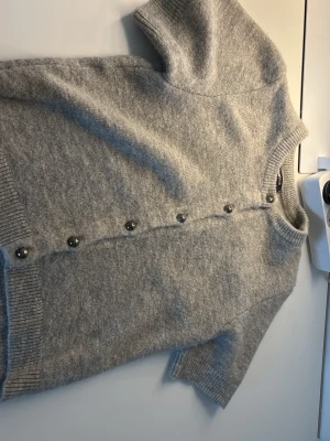 Ljusgrå kortärmad kofta - Mysig ljusgrå kofta med korta ärmar och rund hals. Koftan har ribbade muddar och knäpps framtill med silvriga knappar. Perfekt att slänga över en topp för en avslappnad look. Materialet ser ut att vara mjukt och stickat.