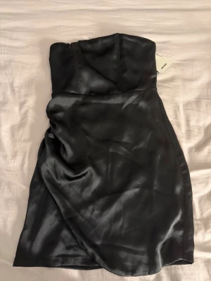 Svart axelbandslös klänning BikBok - Snygg svart axelbandslös kort klänning från BikBok i glansigt silkes  material.  Den är helt ny därav så finns prislappen kvar på klänningen . Klänningen har en figurnära passform och slät yta, perfekt för en trendig look. Enkel men elegant design som passar till många tillfällen som festar . 