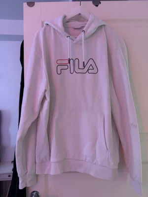Vit hoodie från FILA med logga - Säljer en vit hoodie från FILA med stor logga i svart och rött på bröstet. Hoodien har huva med vita dragsnören och en stor magficka. Tillverkad i mjukt bomullsmaterial och har ribbade muddar vid ärmar och nederkant. Perfekt för en avslappnad och sportig stil.