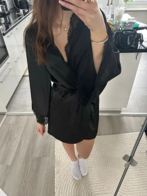 Svart satinmorgonrock med spetsdetaljer - Snygg svart morgonrock i satin med breda spetsdetaljer längs ärmslut och halsringning. Knyts i midjan med matchande band och har en kort, avslappnad passform. 💕