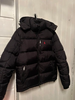 Svart pufferjacka från Polo Ralph Lauren - FIT S. Svart pufferjacka från Polo Ralph Lauren med röd broderad logga på bröstet. Jackan har huva, dragkedja framtill och flera fickor med dragkedja. Quiltad design och klassisk passform, perfekt för kyliga dagar.