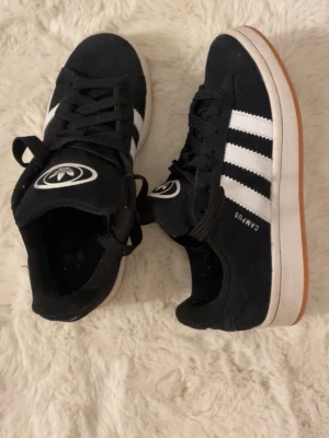 Svarta Adidas Campus sneakers - Säljer ett par klassiska Adidas Campus sneakers i svart mocka med vita ikoniska ränder på sidorna och vit sula. Skorna har svart snörning, Adidas-logga på plösen och hälen samt en brun yttersula. Perfekta för en streetwear-look.
