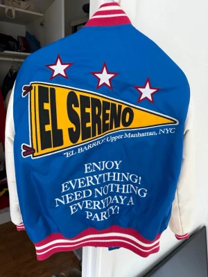 Blå och vit bomberjacka Pull and bear  - Snygg blå och vit bomberjacka från El Sereno med stora tryck och broderade detaljer på både fram- och baksida. Jackan har röda och vita ribbade muddar, tryckknappar och coola patches med text och stjärnor. Perfekt statement-plagg för dig som vill sticka ut.