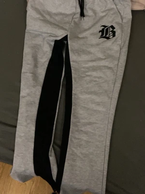 Grå mjukisbyxor med svart detalj - Säljer ett par grå mjukisbyxor med svarta detaljer längs benen och ett tryckt B på vänster sida. Byxorna har resår i midjan med snörning och är tillverkade i mjukt sweatshirtmaterial. Perfekta för chill dagar eller träning.