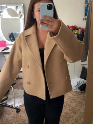 Beige kort kappa  - Stilren beige kappa från vero Moda med dubbelknäppning och bred krage. Modellen är kort och har två snedställda fickor framtill. Perfekt för dig som gillar en trendig och minimalistisk look. Materialet ser ut att vara ullblandning.