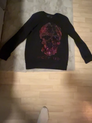 Svart tröja med röd glittrig dödskalle, Philipp Plein  - Cool svart långärmad tröja från Philipp Plein med en stor röd och svart glittrig dödskalle på bröstet. Tröjan har rund hals och diskret logga på ryggen. Perfekt för dig som gillar edgy och unika plagg.