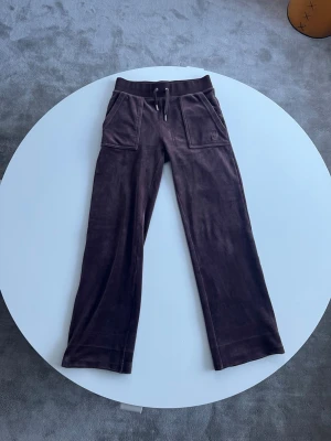 Bruna mjukisbyxor från JUICY COUTURE  - Mysiga bruna velourbyxor från Juicy Couture med broderad logga på fickan, snörning i midjan och fickor både fram och bak. Byxorna har en rak passform. Byxorna är uppsydda så de är kortare än vanligt.💘De är ungefär 65cm från grenen till slutet av benet. 