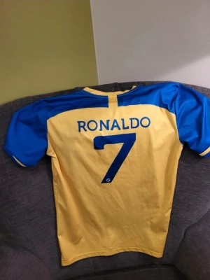 Al Nassr Ronaldo matchtröja gul/blå M - Snygg gul och blå matchtröja från Al Nassr med Ronaldo och nummer 7 på ryggen. Tröjan har klubbens broderade logga på bröstet och är tillverkad i ett lätt och ventilerande material, perfekt för fotboll eller träning.