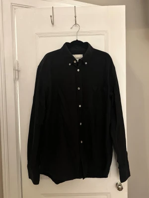 OUR LEGACY svart skjorta - storlek 48/M - Klassisk svart skjorta från OUR LEGACY med button-down krage och vita knappar. Skjortan har lång ärm och en enkel, stilren design som passar till det mesta. Tillverkad i mjuk bomull för skön känsla hela dagen.