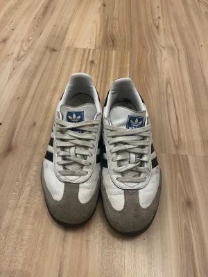 Adidas Samba OG vita och svarta sneakers - Klassiska Adidas Samba OG sneakers i vitt läder med svarta ränder och grå mockadetaljer på tån. Yttersulan är brun och märket syns tydligt på sidan i guld. Skorna har retrovibbar och passar perfekt till streetwear och casual outfits.lite slitna men inget större fel