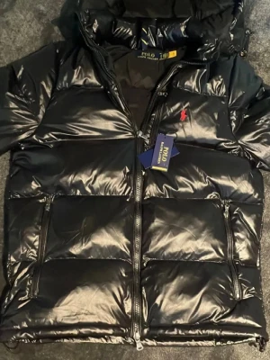 Svart pufferjacka från Polo Ralph Lauren - Säljer en svart, glansig pufferjacka från Polo Ralph Lauren med röd logga på bröstet. Jackan har huva, dragkedja framtill och flera fickor med dragkedjor. Perfekt för kalla dagar och riktigt snygg streetstil.