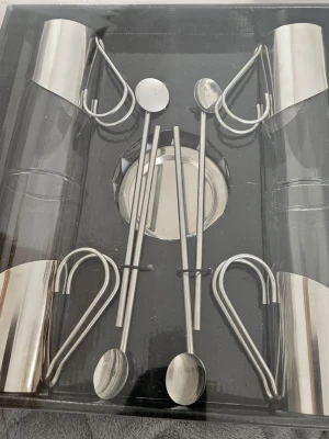 Set med glas och skedar i metall - Säljer ett stilrent set med fyra glas, tillhörande metallhållare, skedar och underlägg. Glasen är cylindriska och hållarna har en unik hjärtformad design i silverfärgad metall. Perfekt för te eller kaffe och ger en modern känsla till köket.