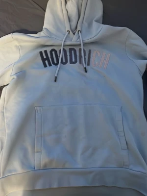 Ljusblå HOODRICH hoodie med tryck - Snygg ljusblå hoodie från HOODRICH med stor magficka och justerbar huva. Framtill står det HOODRICH i svart och vitt tryck. Hoodien har ribbade muddar och är gjord i mjukt bomullsmaterial, perfekt för en avslappnad streetwear-look.