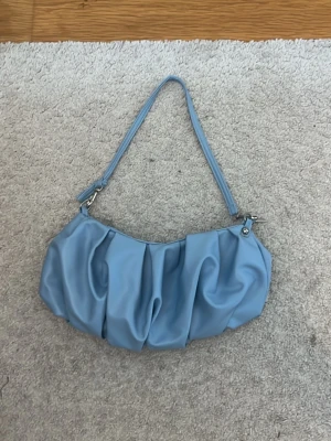 Ljusblå hand väska  - Trendig ljusblå handväska med rynkad design och axelrem. Väskan är gjord i ett slätt fejkläder-material och har silverfärgade metalldetaljer. Perfekt för dig som vill ha en stilren accessoar med lite extra attityd.