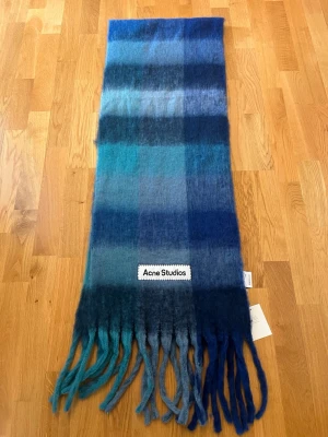 Blå mohairhalsduk från Acne Studios - Säljer en fluffig och mjuk halsduk från Acne Studios i olika blå nyanser med rutigt mönster. Halsduken är tillverkad i mohairblandning och har en stor vävd logotyp. Perfekt accessoar för att lyfta din outfit.