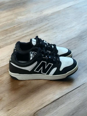 Svarta och vita New Balance sneakers - Snygga sneakers från New Balance i svart och vitt med klassisk N-logga på sidan. Skorna har lågt skaft, perforerad tåbox och svart-vit yttersula. Tillverkade i en mix av läder och syntetmaterial för en clean och sportig look.