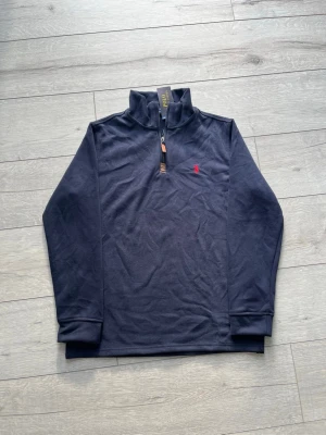 Mörkblå half-zip tröja  - Snygg mörkblå tröja från Ralph Lauren med half-zip och hög krage. Tröjan har en liten röd logga broderad på bröstet och ribbade muddar vid ärmslut och nederkant. Perfekt för en stilren och avslappnad look.