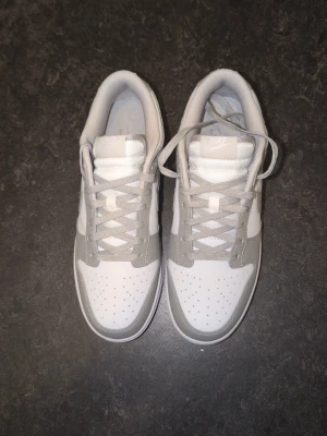 Nike Dunk Low - Snygga Nike Dunk Low sneakers i vitt och ljusgrått läder. Klassisk siluett med perforerad tåbox, vita paneler och grå detaljer på sula, snörning och Swoosh. Perfekta för dig som gillar stilrena och trendiga sneakers.