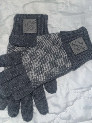 Grå Louis Vuitton stickade handskar - Säljer ett par grå stickade handskar från Louis Vuitton med rutigt mönster och ribbade muddar. Handskarna har en metallbricka med Louis Vuitton Paris-logga på mudden och är perfekta för kalla dagar. Materialet är mjukt och värmande ullblandning.