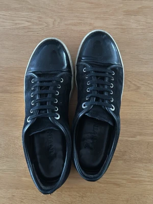 Svarta sneakers från Lanvin - Stilrena svarta sneakers från Lanvin med blank tå i lack och ovandel i mocka. Skorna har vita sulor, svarta skosnören och metallfärgade öljetter. Perfekta för dig som gillar en clean och lyxig look. Storlek 43 dustbags finns