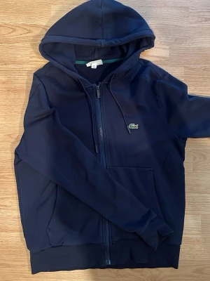 Mörkblå hoodie från Lacoste - Snygg mörkblå hoodie från Lacoste med dragkedja framtill och klassisk krokodillogga på bröstet. Hoodien har huva med dragsko och praktiska fickor på magen. Perfekt för en avslappnad och sportig stil. Materialet känns mjukt och bekvämt.
