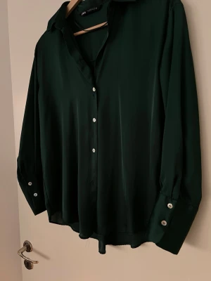 Mörkgrön skjorta från Zara - Stilren mörkgrön skjorta från zara med klassisk krage och knäppning framtill. Den har långa ärmar med breda manschetter och silvriga knappar. Tillverkad i ett mjukt, lätt och glansigt material som ger en elegant känsla.