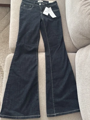 Mörkblå bootcut jeans med kontrastsömmar - Snygga mörkblå bootcut jeans med vita kontrastsömmar och klassisk femficksmodell. Jeansen har hög midja och vida ben, perfekta för en trendig och avslappnad look. Tillverkade i stretchig denim för extra komfort.