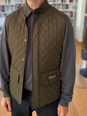 Olivgrön väst Belstaff - Snygg olivgrön quiltad dunväst från Belstaff med hög krage och tryckknappar. Västen har två stora fickor framtill med lock och Belstaff-logga på ena fickan. Perfekt för lager-på-lager och ger en stilren look. Aldrig använd, du kan köpa denna och belstaff jackan för ett bättre pris 😁