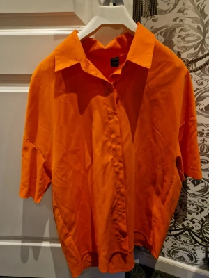 Orange kortärmad skjorta - Säljer en orange skjorta med korta ärmar och klassisk krage. Skjortan har knappar framtill och är tillverkad i ett mjukt tyg som känns skönt mot huden. Perfekt för dig som vill sticka ut med färg i din outfit.