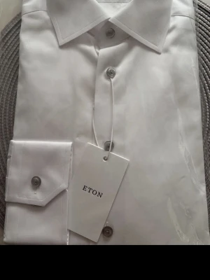 Vit skjorta från Eton - Klassisk vit skjorta från Eton med knappar framtill och elegant krage. Tillverkad i ett slätt och exklusivt material som ger en stilren look. Perfekt för dig som vill ha en tidlös och snygg skjorta i garderoben.