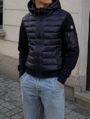 Marine blue Moncler Cardigan med NFC - Snygg lila pufferjacka från Moncler med huva och stickade ärmar. Jackan har dragkedja framtill, två fickor och en extra ficka med dragkedja på ärmen. Moncler-logga på armen och ribbade muddar vid ärmslut och nederkant. Perfekt för kalla vinterdagar.