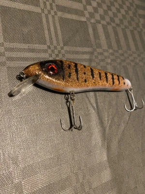 Abu garcia Svartzonker - Säljer ett fiskedrag med glittrig brun och svart randig design, röda ögon och två trekrokar. Draget har en genomskinlig sked framtill och är perfekt för fiske efter rovfisk. Snygg och detaljerad finish. Pris kan diskuteras 