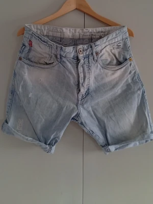 Ljusblå jeans shorts  - Säljer ett par ljusblå denimshorts från Jack & Jones med klassisk femficksdesign och uppvikta benslut. Shortsen har normal passform och detaljerade sömmar på bakfickorna. Perfekta för varma dagar och enkel att matcha med olika stilar.