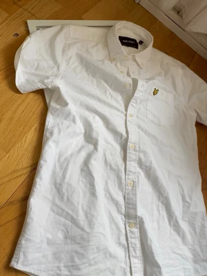 Vit kortärmad skjorta Lyle & Scott - Klassisk vit skjorta från Lyle & Scott med korta ärmar och knappstängning framtill. Bröstficka med gul logga i form av en fågel. Skjortan är i bomull och har en avslappnad passform, perfekt till sommaren.