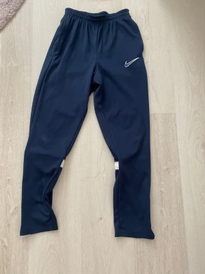 mörkblåa nike träningsbyxor - Snygga mörkblå träningsbyxor från Nike med vit logga på ena låret. Byxorna har resår i midjan, smal passform och vita detaljer baktill vid vaderna. Perfekta för träning eller sportaktiviteter. Materialet är troligtvis polyester för bra rörelsefrihet.