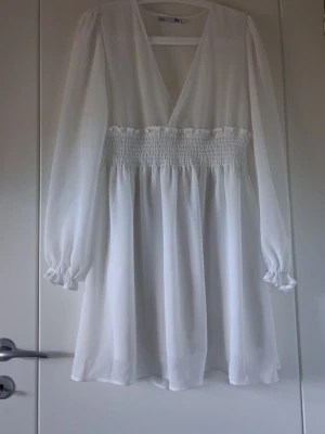 Vit kortklänning med v-ringning - Säljer en vit kortklänning med v-ringning och långa ärmar. Klänningen har smock i midjan som ger en snygg siluett och är tillverkad i ett lätt, flödande material. Perfekt för sommarens alla tillfällen!