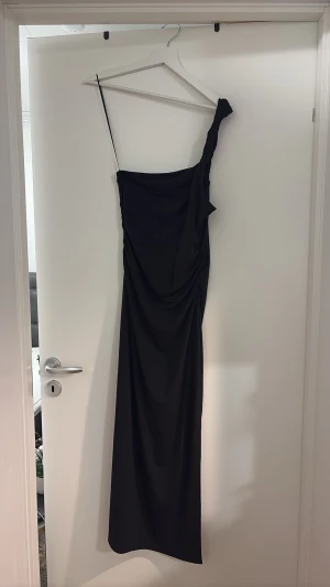 Svart oneshoulder långklänning - Säljer en elegant svart långklänning med oneshoulder-design och draperad detalj längs sidan. Klänningen har en smal passform och är tillverkad i ett mjukt, följsamt material som ger en snygg siluett. Perfekt för dig som vill sticka ut med en stilren look.