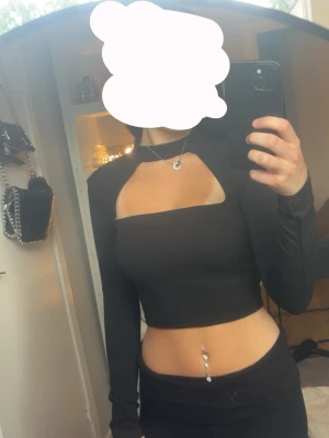 Svart croppad topp från Pull&Bear - Trendig svart croppad topp från Pull&Bear med lång ärm och unik cut-out detalj vid halsen. Toppen har en tight passform och är perfekt för dig som vill sticka ut. Tillverkad i mjukt syntetmaterial som sitter skönt mot kroppen.