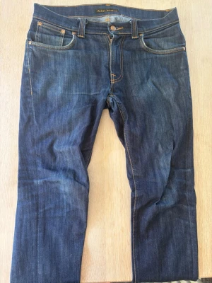 Mörkblå jeans från Nudie Jeans - Säljer ett par mörkblå jeans från Nudie Jeans i modellen thinn fin med klassisk femficksmodell och kontrastsömmar. Jeansen har raka ben och snygga slitningar samt Nudie-logga på bakfickorna. Tillverkade i slitstarkt denimtyg. Hör gärna av er vid frågor!